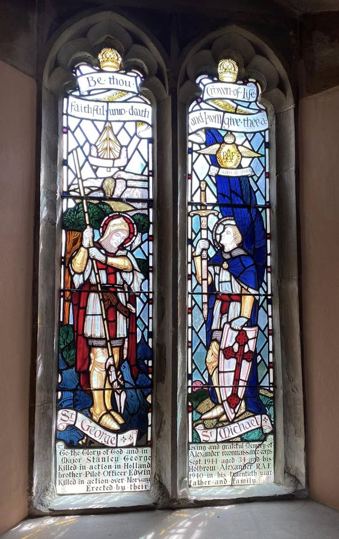 Stained glass memorial window featuring two panels: one showing St George with a red cross shield and spear, and the other St Michael with blue wings and a sword. Below is a dedication to Major Stanley Alexander, killed in action in Holland in 1944, and his brother Pilot Officer Edwin Alexander, killed in action over Norway in 1940