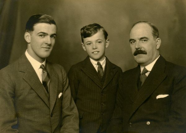 Sepia-toned studio portrait of three males from different generations, dressed in suits. A young boy stands between an older man with a moustache and a younger man, depicting a family group of father, son, and younger brother